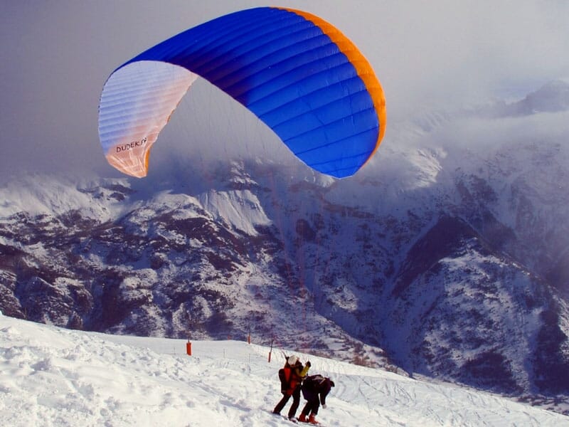 Parapente biplace s'apprêtant à décoller sur une pente enneigée, avec un parachute bleu et orange dans un ciel nuageux et un paysage montagneux en arrière-plan