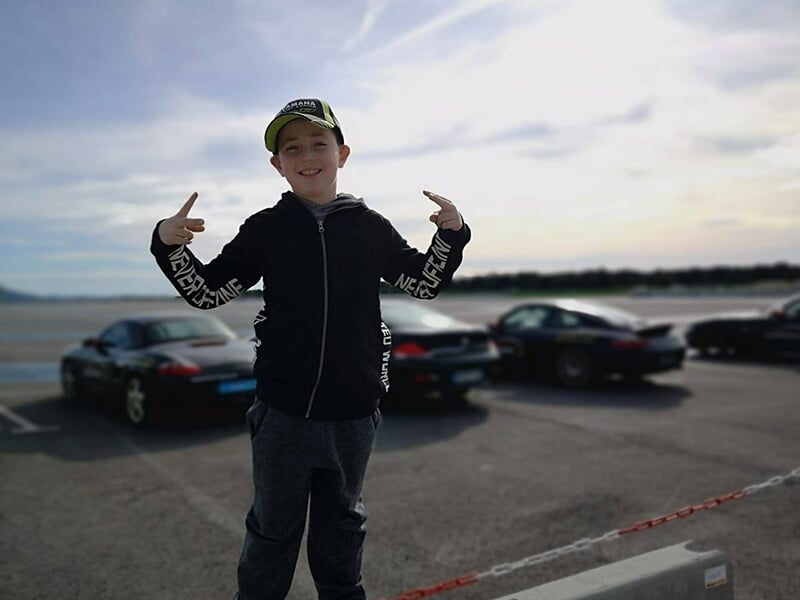 Un enfant portant une casquette pose devant des voitures de sport sur un circuit du Var