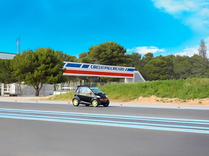 Petite voiture électrique noire avec détails orange et jantes vert fluo roulant sur le circuit Paul Ricard, avec un bâtiment emblématique en arrière-plan et des lignes bleues sur la piste