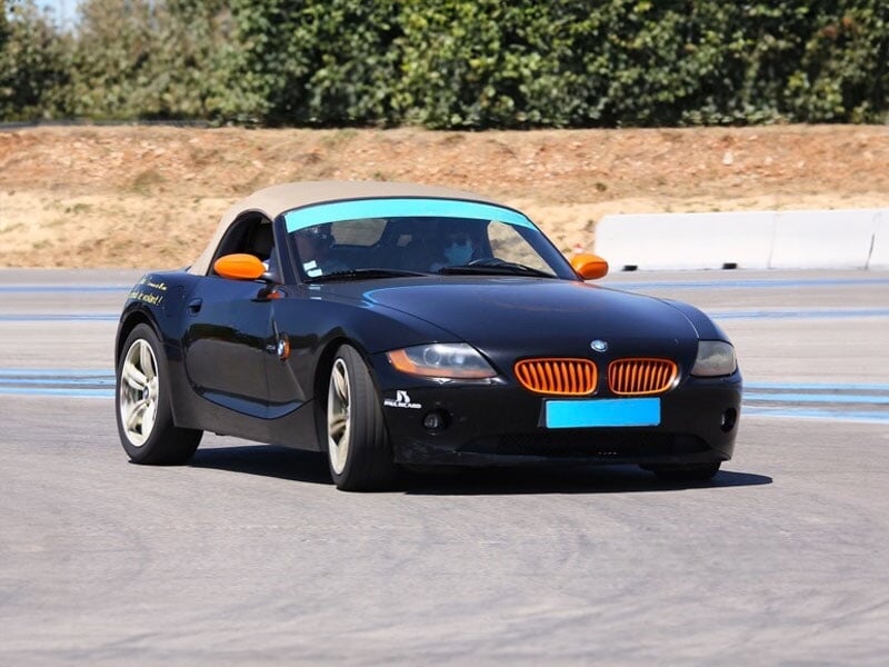 Roadster noir avec capote beige et calandre orange roulant sur une piste asphaltée, entourée de verdure et d’une zone dédiée au pilotage