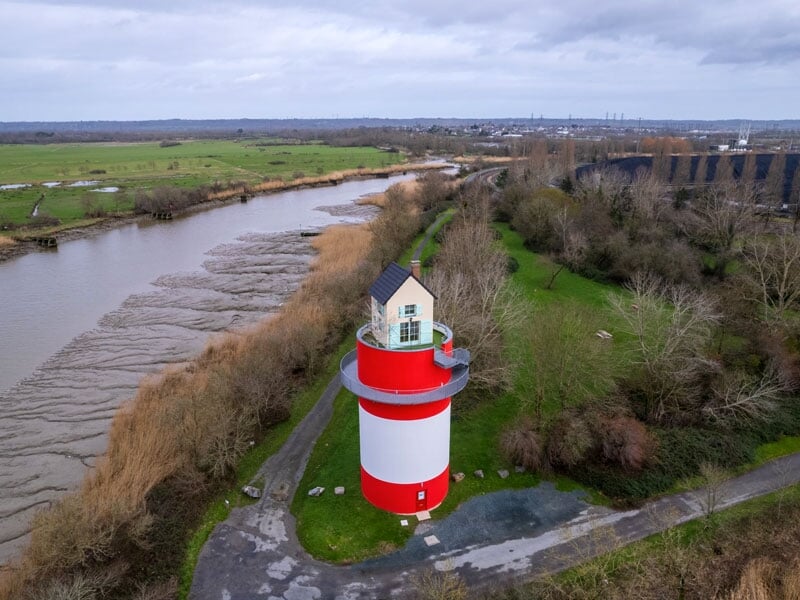 Phare rouge et blanc, style classique, sur une petite élévation près d'un fleuve boueux avec des arbres nus et une digue en arrière-plan, dans une atmosphère hivernale