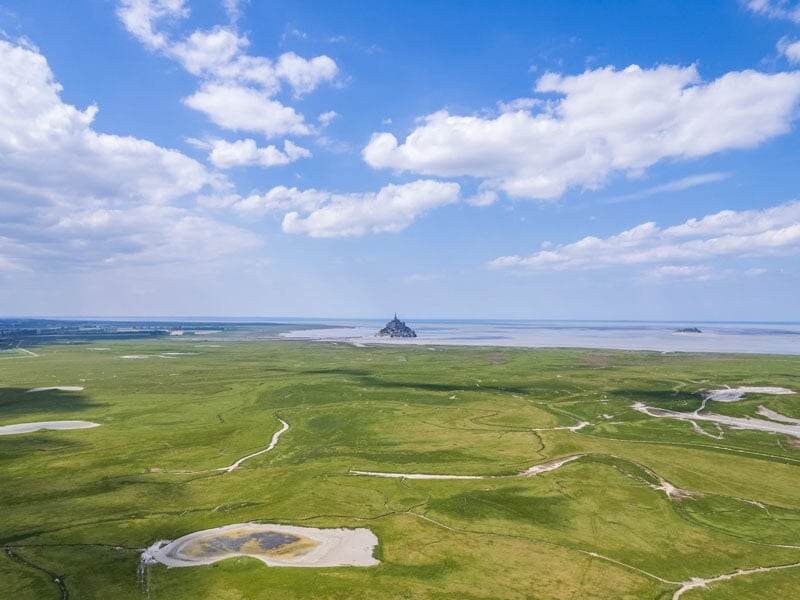 Paysage marécageux vaste avec des cours d'eau sinueux et une végétation verdoyante, dominé au loin par le Mont Saint-Michel sous un ciel bleu parsemé de nuages