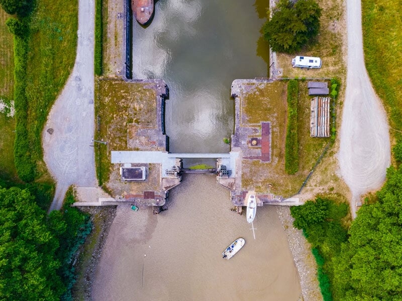 Vue aérienne d'une écluse avec une petite embarcation blanche amarrée, encadrée par la verdure des berges et deux chemins de terre, sous un ciel nuageux