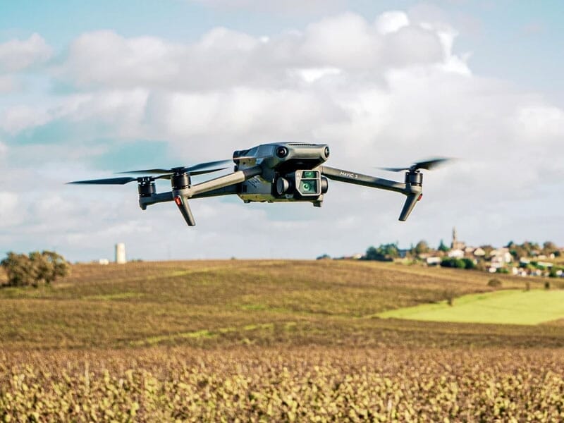 Drone quadricoptère en vol au-dessus d'un champ labouré avec horizon rural et ciel nuageux en arrière-plan, mettant en avant la technologie de surveillance et de photographie aérienne