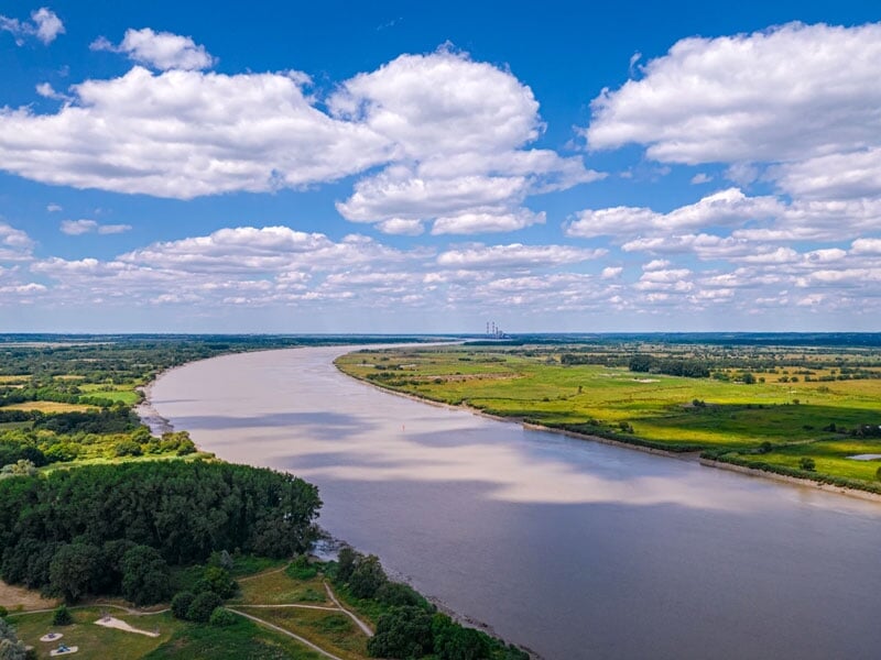 Panorama fluvial capturé depuis les airs, montrant un large fleuve serpentant à travers une plaine verdoyante, sous un ciel parsemé de nuages blancs volumineux