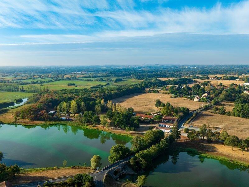 Vue aérienne d'un paysage rural avec des plans d'eau, des champs verts et des routes sous un ciel bleu clair.