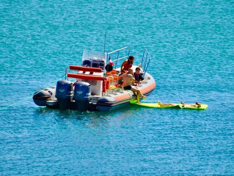 Bateau pneumatique multicolore sur une eau turquoise, équipé de trois moteurs hors-bord bleus, transportant plusieurs personnes, certaines pêchant à l'arrière, d'autres assises à l'avant sur un matela