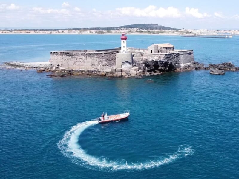 Vue aérienne d'un bateau rouge créant un sillage circulaire sur l'eau bleue près d'un vieux fort avec un phare rouge et blanc, situé sur un îlot rocheux entouré par la mer sous un ciel clair.