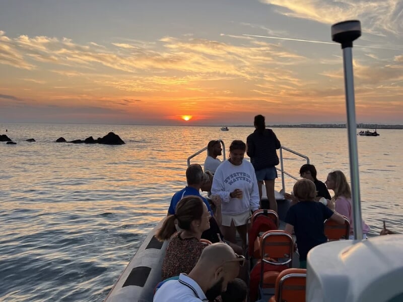 Excursion en mer au coucher du soleil avec des passagers à bord d'un bateau pneumatique observant le soleil qui se couche à l'horizon, créant un ciel teinté de nuances orange et rose, reflétant sur l'