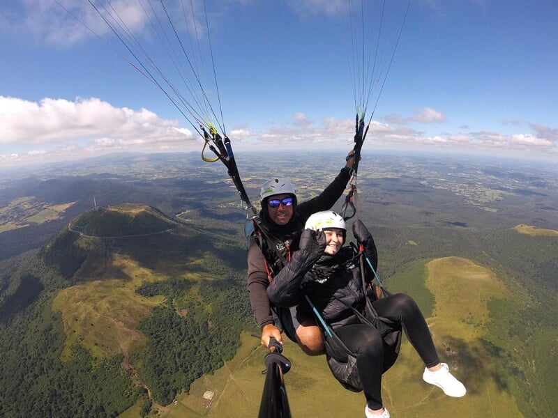 Parapentiste en tandem et son passager volant haut dans le ciel, avec une vue panoramique sur des collines, des forêts et des champs