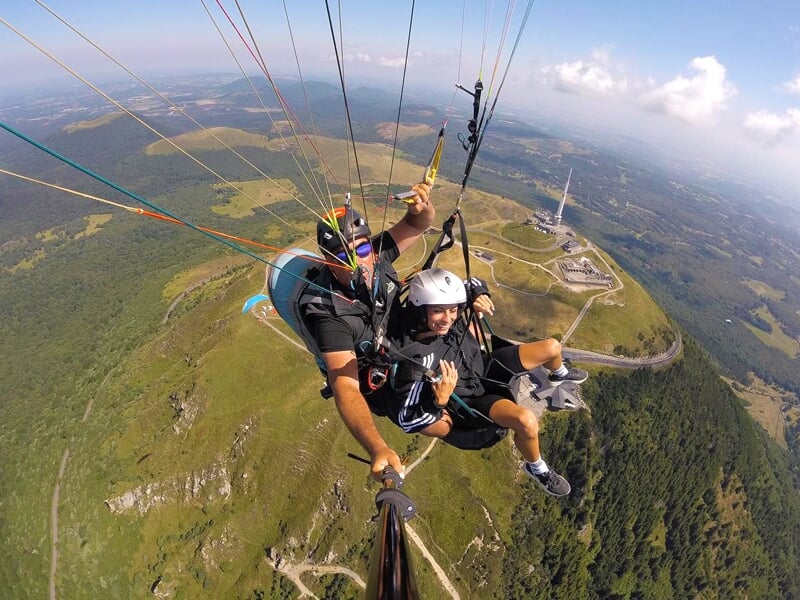 Deux personnes en tandem de parapente sourit à la caméra, avec une vue aérienne de collines verdoyantes et de routes en arrière-plan