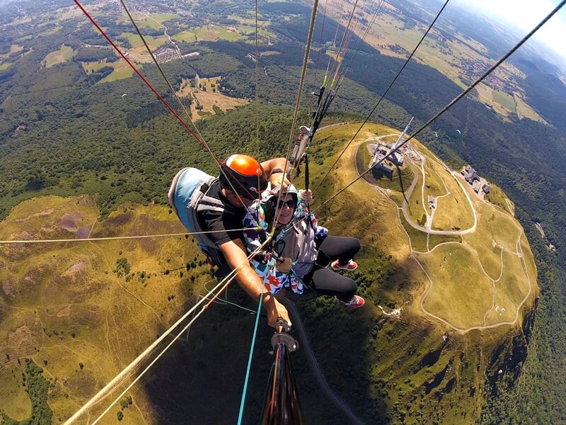 Deux personnes en tandem de parapente survolent un paysage vallonné avec des champs et des forêts, vue du dessus