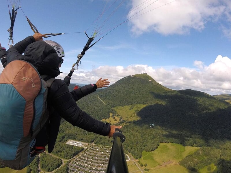 Parapentiste en combinaison noire étendant la main vers des collines verdoyantes et des parapentes en distance, ciel nuageux en arrière-plan