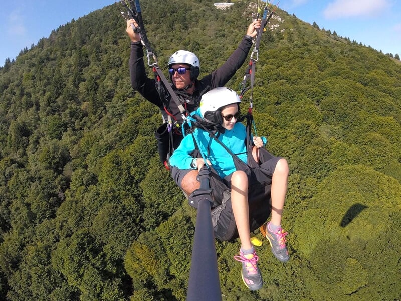 Parapentiste et passager en tandem survolent une dense forêt de conifères, vue latérale avec des montagnes en arrière-plan