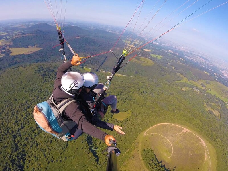 Vue de dos d'un parapentiste en vol solo au-dessus d'une forêt et d'une plaine, avec une grande ombre du parapente sur le sol