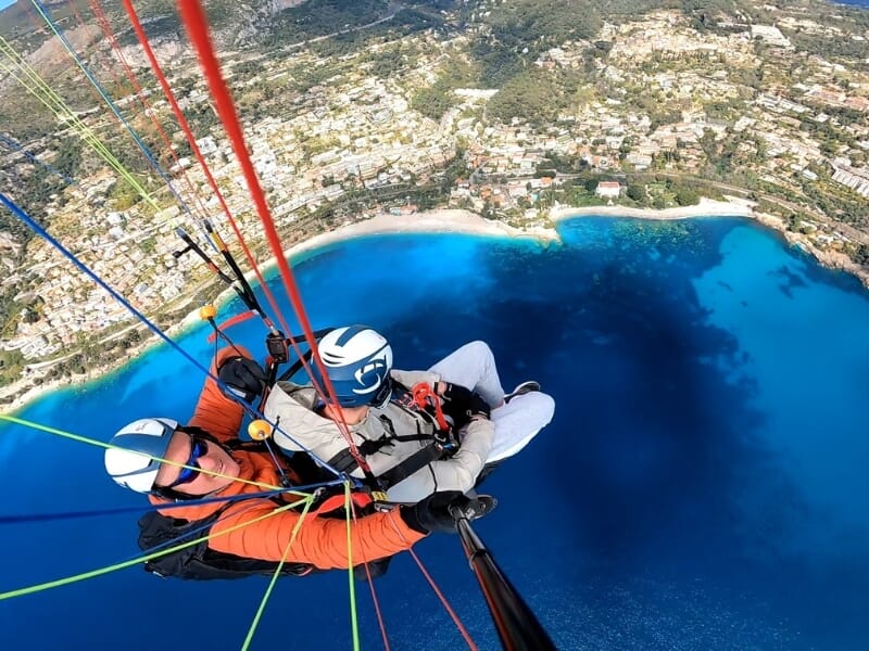 Vue en parapente en tandem au-dessus d'un paysage côtier, avec un passager et un instructeur en vol au-dessus d'une eau bleu turquoise et d'une ville côtière.