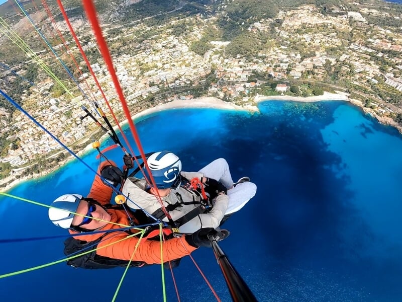Vue en parapente en tandem au-dessus d'un paysage côtier, avec un passager et un instructeur en vol au-dessus d'une eau bleu turquoise et d'une ville côtière.