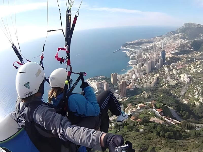 Deux personnes en vol en parapente avec vue sur une ville côtière et la mer, les deux portant des casques et attachées au harnais de parapente.