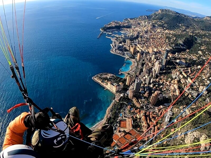 Vue en altitude de deux parapentistes en tandem survolant une zone urbaine côtière avec des routes sinueuses et un paysage urbain dense en dessous.