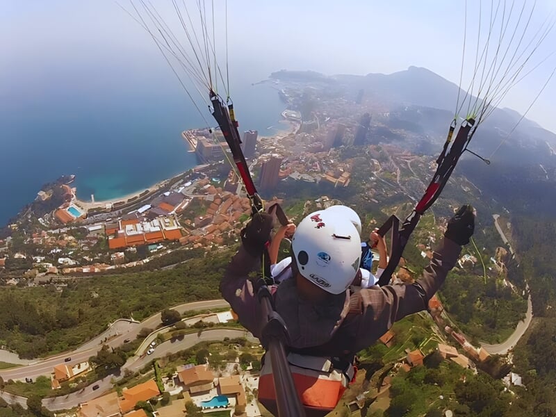 Parapentistes en tandem s'élevant au-dessus d'une ville côtière avec vue sur la mer Méditerranée, des bâtiments urbains et des montagnes en arrière-plan.