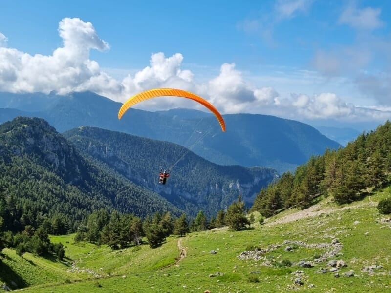 Un parapente orange vole au-dessus d'une vallée verdoyante et montagneuse sous un ciel bleu parsemé de nuages.