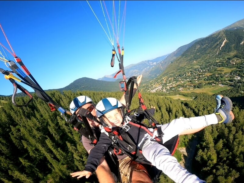 Deux personnes en parapente en tandem, une devant l'autre, survolent un paysage forestier avec des montagnes à l'horizon sous un ciel bleu.
