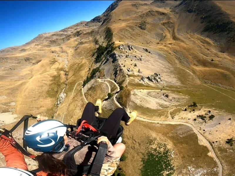Vue en perspective de la première personne d'un parapente en tandem survolant un paysage montagneux sec et rocheux, avec des chemins sinueux en dessous.
