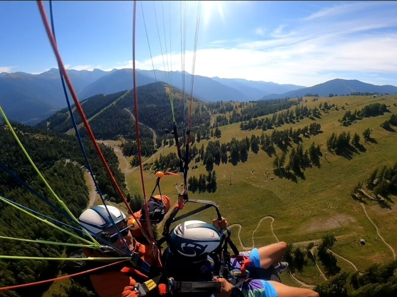 Parapentistes en tandem survolant une vallée montagneuse verdoyante, avec des lignes de parapente colorées s'étendant vers le haut de la photo.