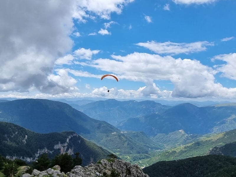 n parapente orange s'élevant au-dessus d'une chaîne de montagnes verdoyantes et de vallées avec des nuages ??blancs flottant dans le ciel bleu au-dessus.