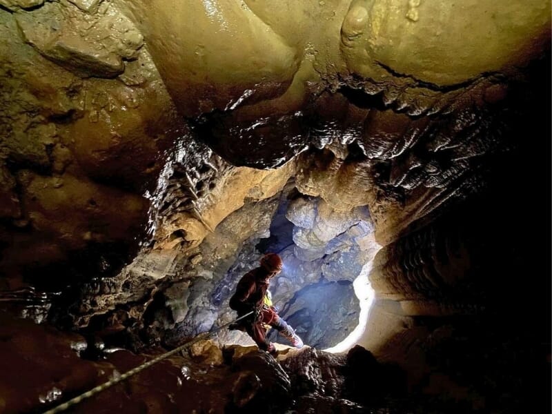 Spéléologue explorant une grotte vaste avec des formations rocheuses et stalactites éclairées par la lumière naturelle venant d'une ouverture dans la caverne