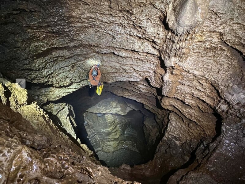 Vue plongeante sur un spéléologue debout sur un rocher dans une grotte profonde avec un casque et une lampe frontale, prêt pour la descente.