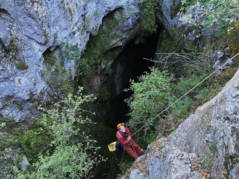 Spéléologue en combinaison rouge et casque descendant une corde à côté d'une grande entrée de caverne entourée de rochers et de végétation