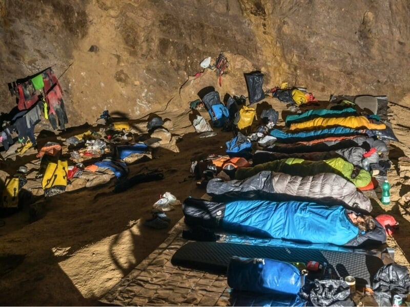 Camp de base temporaire dans une grande caverne, avec des spéléologues reposant dans des sacs de couchage colorés et de l'équipement éparpillé tout autour sur le sol de la grotte
