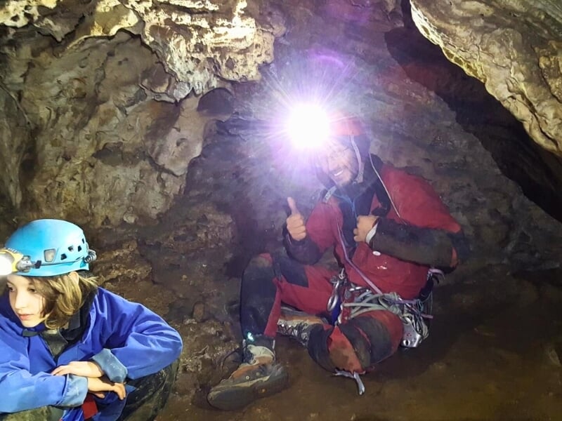 Un spéléologue souriant avec un enfant à ses côtés à l'intérieur d'une grotte, avec une lumière brillante provenant de sa lampe frontale