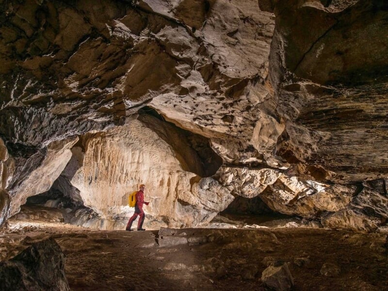 Une personne marchant dans une grande caverne éclairée, soulignant les textures riches et variées de la roche, avec un sac à dos rouge