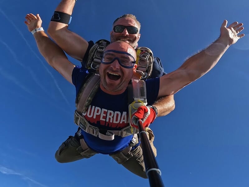 Deux parachutistes en chute libre, bras ouverts et visages rayonnants sous un ciel dégagé.