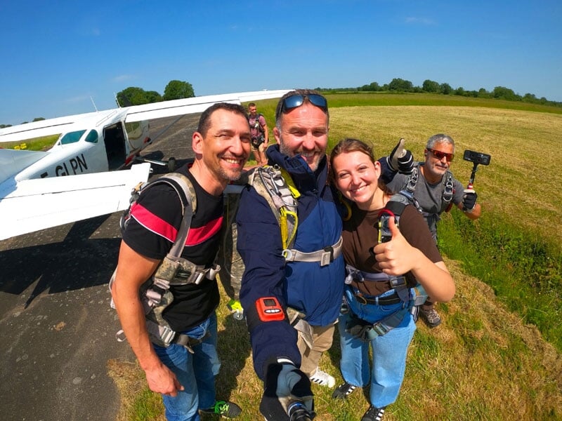 Groupe de parachutistes souriants posant près d'un avion sur une piste, avec harnais de sécurité et ciel bleu dégagé. Activité de parachutisme en plein air