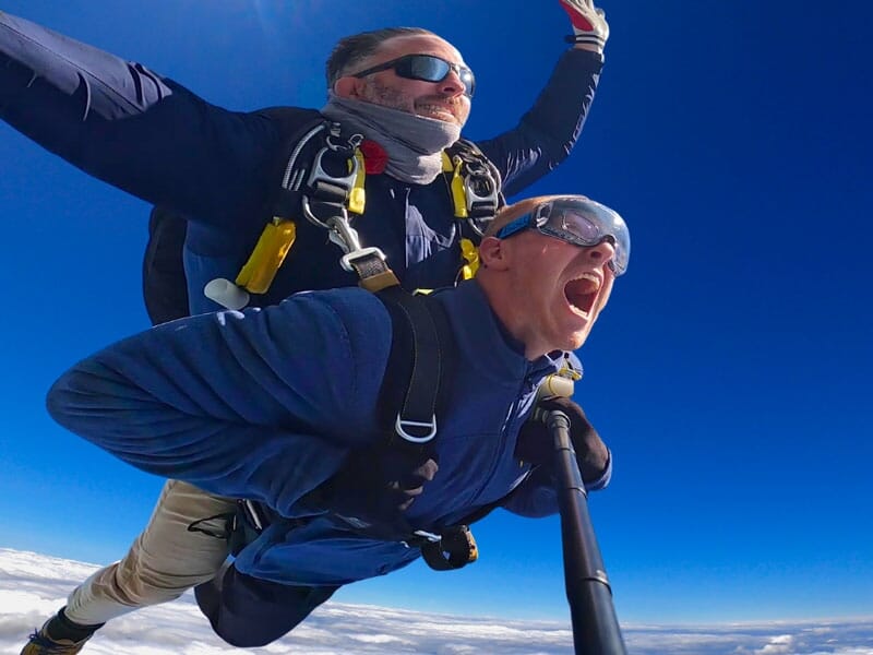 Tandem de parachutisme en chute libre au-dessus des nuages, expressions de joie et d'excitation, ciel bleu éclatant. Aventure aérienne extrême