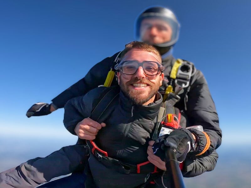 Parachutiste en tandem souriant avec lunettes de protection et harnais, fond flou d'un ciel dégagé, moment capturé en plein saut. Sensations fortes garanties
