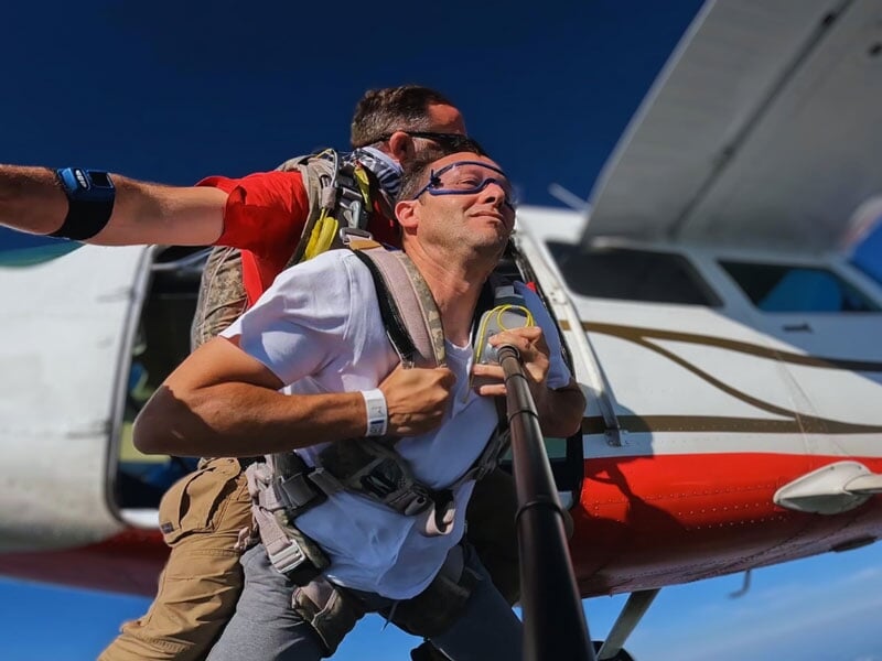 Parachutiste en tandem sautant d'un avion, harnais attachés et instructeur derrière. Avion blanc avec détails rouges visible, ciel dégagé. Départ d'une expérience en parachute