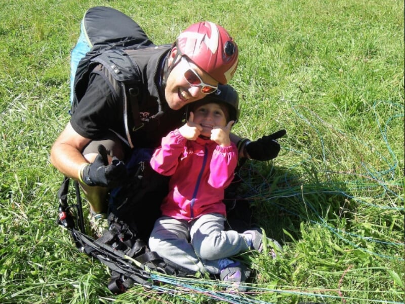 Homme en casque rouge et lunettes de soleil se repose dans une prairie avec une petite fille en veste rose, qui fait un signe de