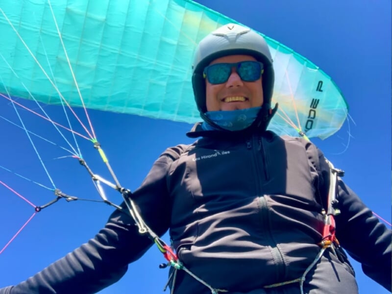 Homme souriant sous un parapente, équipé d'un casque noir et de lunettes de soleil teintées bleues, porte une veste noire. Le ciel bleu clair en arrière-plan met en valeur la voile turquoise du parape