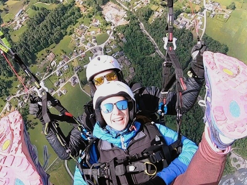 Vue aérienne lors d'un vol en tandem en parapente, montrant deux personnes souriantes équipées de casques et de lunettes de soleil. L'une porte une combinaison noire et bleue, et l'autre une veste noi