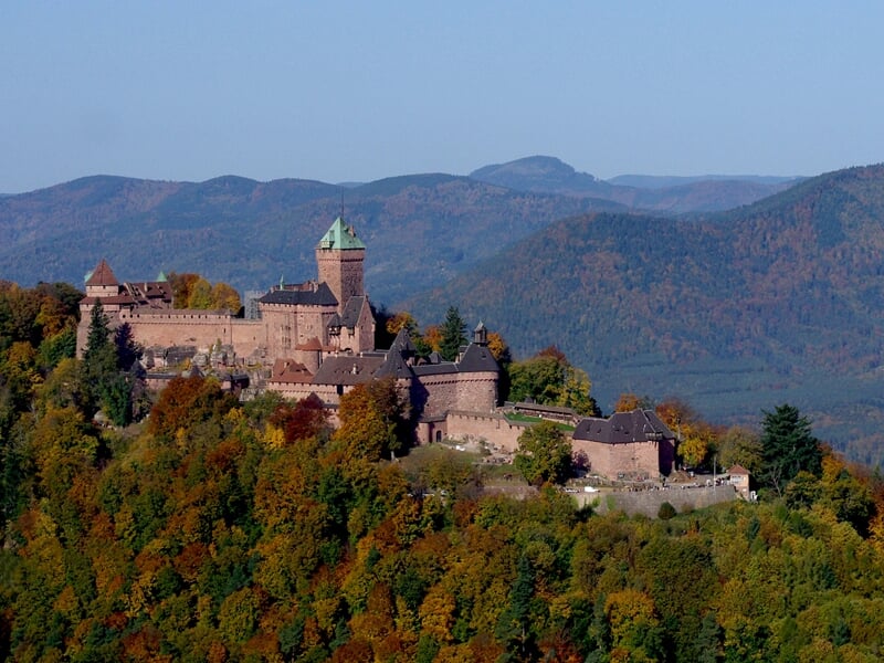 Vue aérienne d'un château médiéval entouré de forêts aux couleurs d'automne sous un ciel bleu clair.