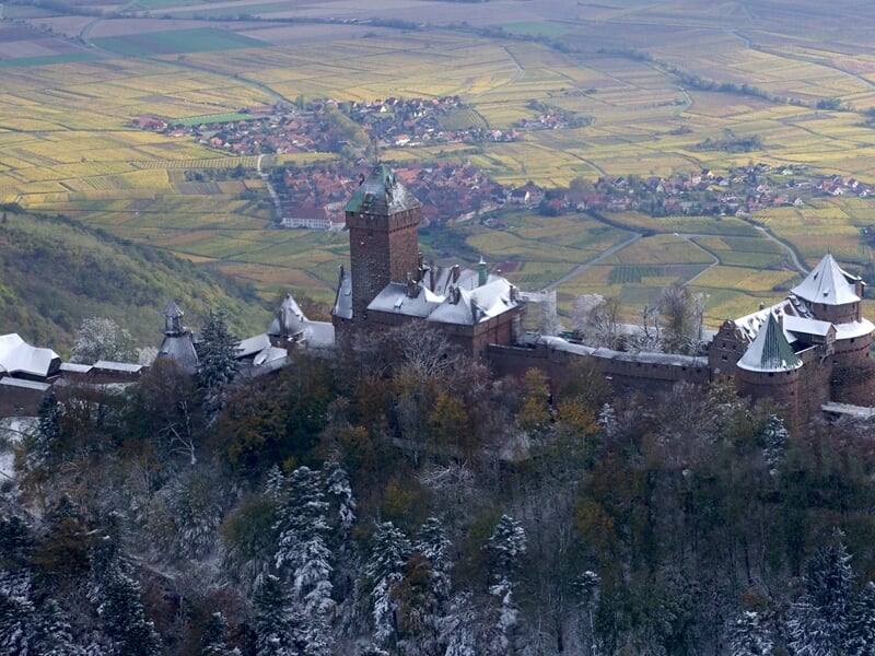 Château fortifié sur une colline enneigée avec un village et des champs en arrière-plan sous un ciel clair.