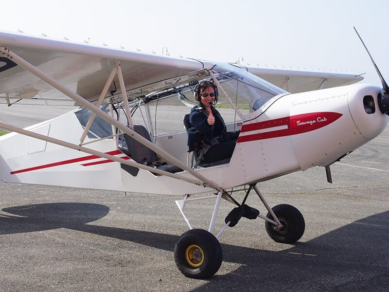 Pilote assis dans un petit avion blanc et rouge sur une piste, prêt à décoller ou après l'atterrissage.