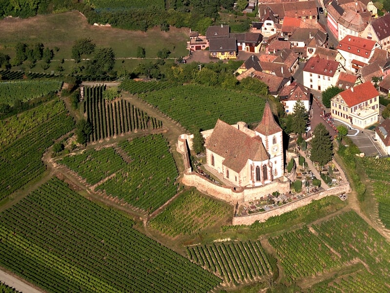 Vue aérienne d'une petite église entourée de vignes et de collines verdoyantes sous un ciel clair.