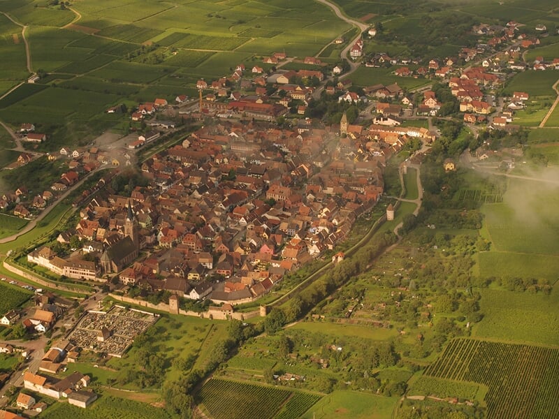 Vue aérienne d'un village fortifié entouré de champs verts et de collines sous un ciel partiellement nuageux.