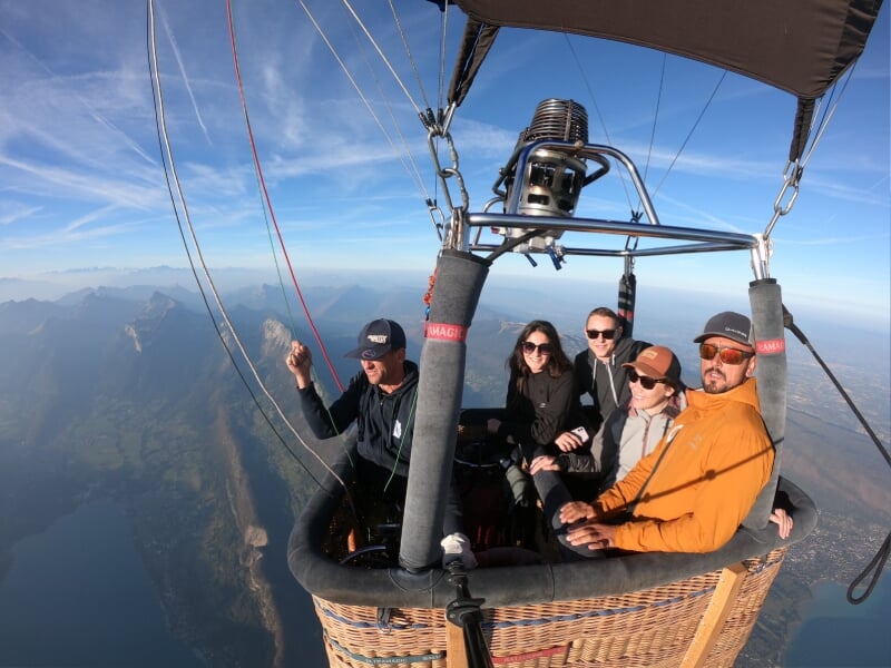 Groupe de personnes profitant d'un vol en montgolfière, survolant un paysage de montagnes et de lacs par une journée ensoleillée avec un ciel bleu dégagé.