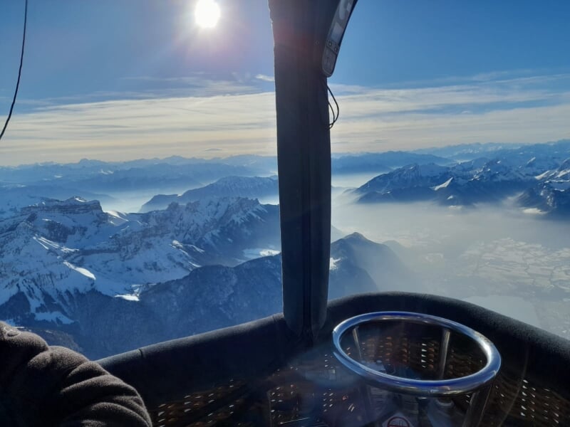 Lever de soleil sur une chaîne de montagnes enneigées, observé depuis une montgolfière, avec un ciel clair et brumeux au-dessus des vallées.
